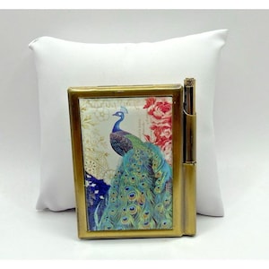 May include: A gold-coloured metal cigarette case with a peacock design. The case is rectangular with a hinged lid and a built-in lighter. The peacock has blue, green, and gold feathers against a floral background. The case is approximately 9 cm in height.
