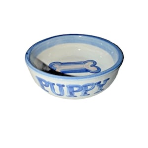Puede incluir: Un cuenco de cerámica blanca para perros con borde azul y la palabra "PUPPY" en letras azules. El cuenco presenta un diseño en forma de hueso azul y blanco en el centro, ideal para un perro pequeño o cachorro.