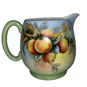 May include: A vintage porcelain pitcher with a blue and green floral design featuring apples and leaves. The pitcher has a handle and a rounded body.