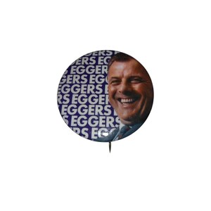 May include: A circular pinback button featuring a photographic portrait of a smiling person in a suit and tie. The background consists of the repeated text EGGERS in white capital letters against a dark blue field. A metal pin is visible at the bottom.