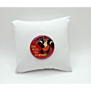 Op de afbeelding: Een ronde, rode knoop met een cartoonafbeelding van een zwart-witte koe met een rood vest. De knoop heeft de tekst "Get with the program!" in geel en wit. De knoop ligt op een wit kussen.