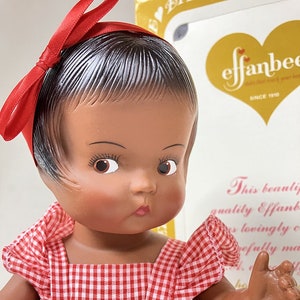 Könnte beinhalten: Eine Puppe mit brauner Haut und braunen Augen, die ein rot-weiß kariertes Kleid und ein rotes Schleifenband trägt. Die Puppe hält ihre rechte Hand hoch. Die Puppe steht vor einer Schachtel mit dem Text "effanbee since 1910".
