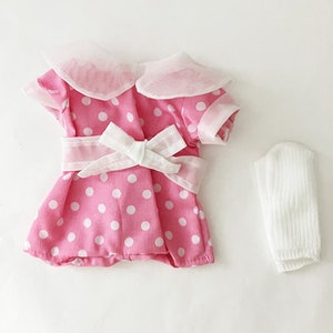 Può includere: Un vestito a pois rosa e bianco con un fiocco bianco e un colletto bianco. Il vestito ha maniche corte e un calzino bianco è incluso.