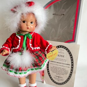Puede incluir: Muñeca Patsy vintage con un atuendo festivo rojo y verde con detalles de plumas blancas. La muñeca tiene un sombrero de plumas blancas y zapatos rojos. Se incluye un certificado de autenticidad.