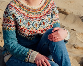 Può includere: Un maglione fantasia in verde acqua, crema e rosso con maniche lunghe. Il maglione presenta uno scollo tondo e un design geometrico dettagliato sul petto e sulle spalle. La persona indossa jeans blu e scarpe da ginnastica bianche seduta sulla sabbia.