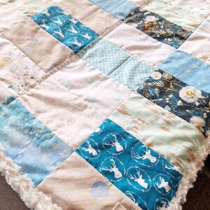 May include: A patchwork quilt with a variety of fabrics in shades of blue, beige, and white. Some squares feature deer silhouettes, floral patterns, and polka dots. The quilt has a soft, white, fringed edge.