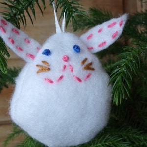 May include: White felt bunny ornament with pink and gold embroidered details. The bunny has blue eyes and a pink nose.