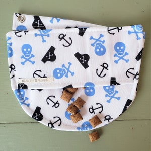 May include: A white fabric dog bandana with a blue and black skull and crossbones, anchor, and pirate hat pattern. The bandana has a fold-over flap and the text "La Rue de Fleurs" is printed on the flap.