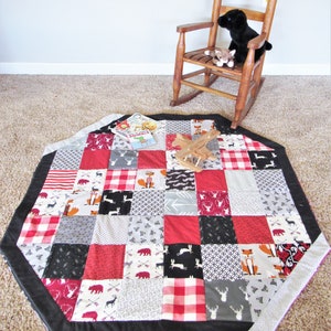 May include: A red, black, and white patchwork playmat with a black border. The playmat is shaped like an octagon and has a variety of patterns, including plaid, polka dots, and animal prints. A wooden toy airplane and a book titled "Animals" are on the playmat.