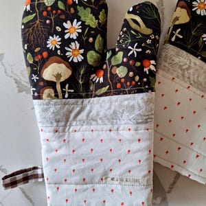 Peut inclure: Un ensemble de manique et de dessous de plat. Le gant de cuisine présente un fond noir avec un motif floral et de champignons. La partie inférieure est blanche avec de petits motifs floraux rouges. Le dessous de plat a un design similaire. Une boucle à carreaux marron et blanc est attachée.