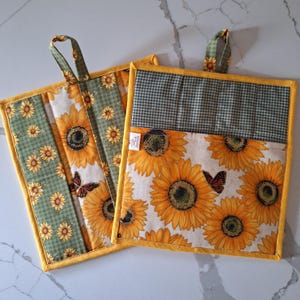 May include: Two square pot holders with a sunflower and butterfly pattern. The pot holders have a green and yellow gingham pattern on the top and a yellow border. Each pot holder has a fabric loop for hanging.