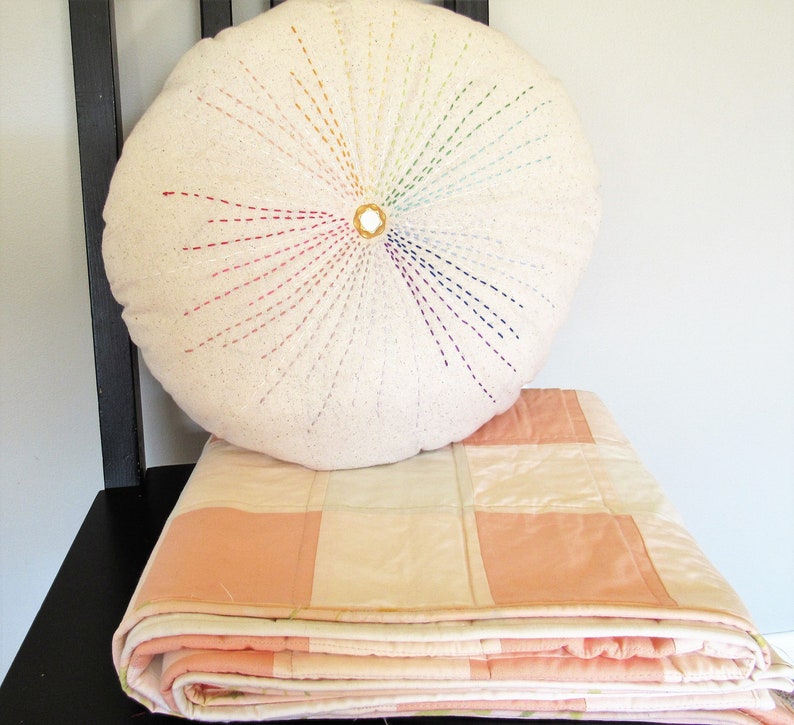 May include: A white round pillow with a rainbow-colored embroidery design in the center. The pillow is sitting on top of a folded pink and white quilt.