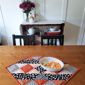 May include: A diamond-shaped Halloween table runner with a patchwork design featuring black, orange, and white fabric. The runner has a variety of patterns including ghosts, pumpkins, and polka dots. A white bowl filled with yellow candy corn sits on the runner.