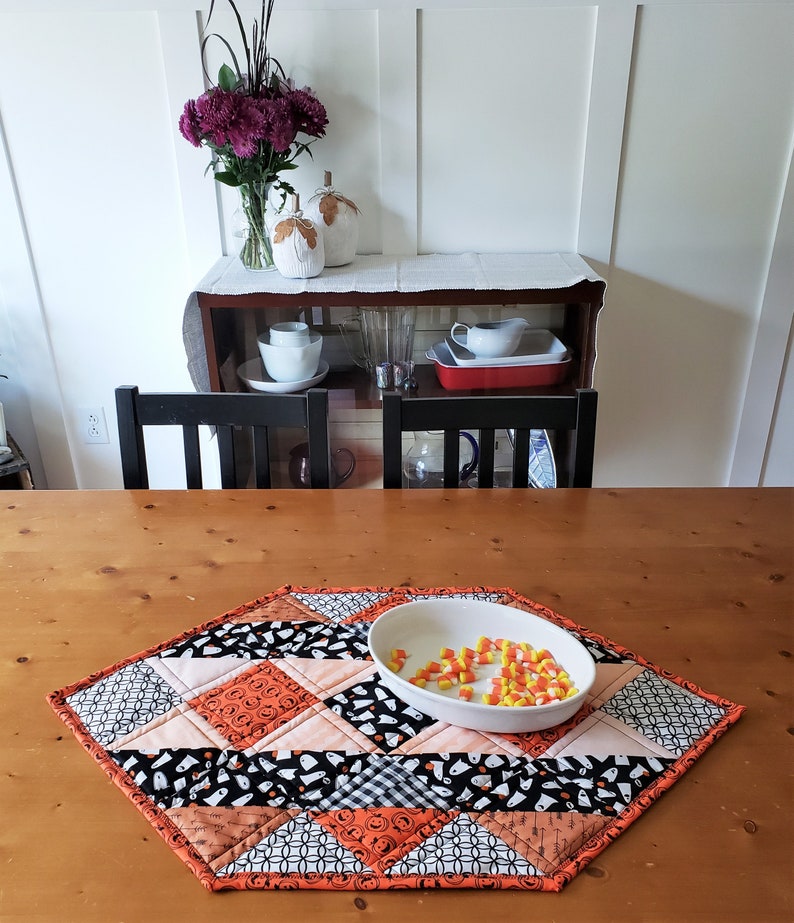 May include: A diamond-shaped Halloween table runner with a patchwork design featuring black, orange, and white fabric. The runner has a variety of patterns including ghosts, pumpkins, and polka dots. A white bowl filled with yellow candy corn sits on the runner.