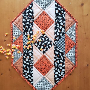 May include: A diamond-shaped table runner with a Halloween theme. The runner is made of patchwork fabric with various patterns, including ghosts, pumpkins, and black and white checkered patterns. The runner is trimmed with a black and orange border. Candy corn is scattered on a wooden surface next to the runner.