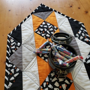 May include: A black, white, and orange quilted table runner with a ghost pattern. A black ceramic jar filled with colorful candy is on top of the runner.
