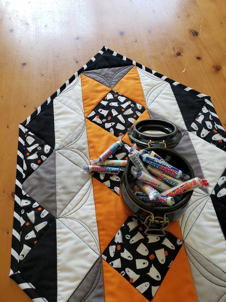 May include: A black, white, and orange quilted table runner with a ghost pattern. A black ceramic jar filled with colorful candy is on top of the runner.