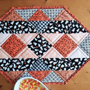 May include: A diamond-shaped Halloween table runner with a black and white geometric pattern, orange and black pumpkin faces, and black and white ghost patterns. The runner is trimmed with orange fabric.