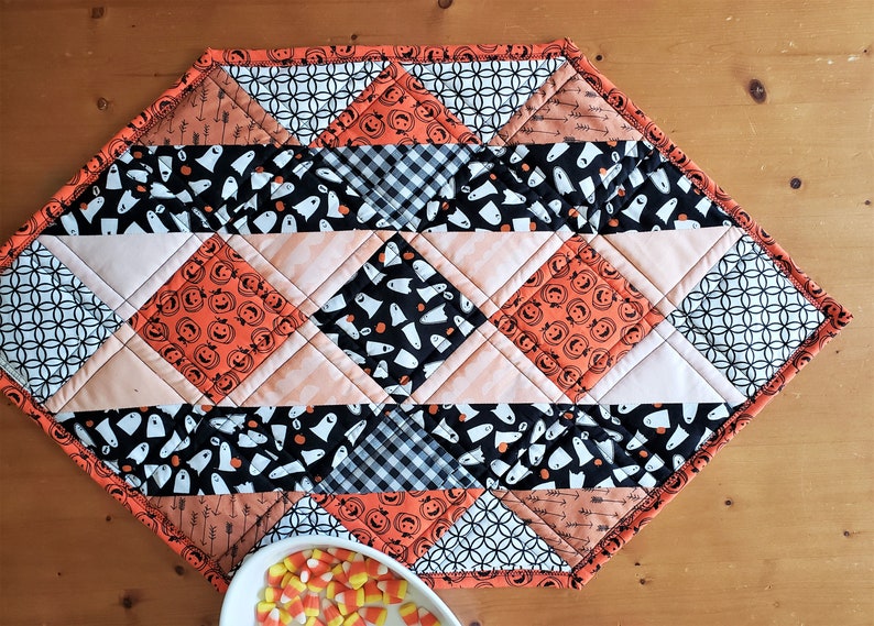 May include: A diamond-shaped Halloween table runner with a black and white geometric pattern, orange and black pumpkin faces, and black and white ghost patterns. The runner is trimmed with orange fabric.