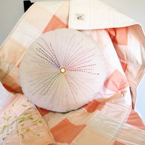 May include: A round, white pillow with colorful embroidery in the shape of a sunburst. The pillow is sitting on a pink and white patchwork quilt.