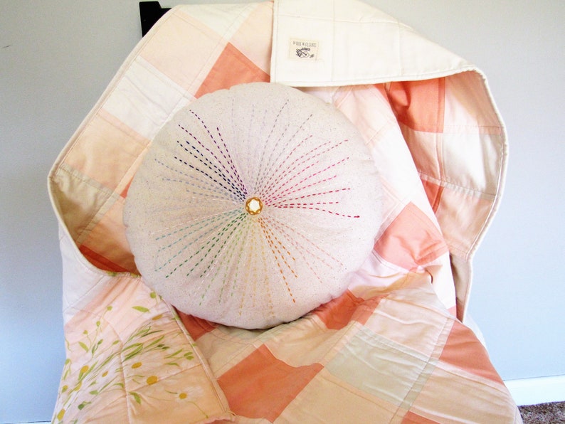 May include: A round, white pillow with colorful embroidery in the shape of a sunburst. The pillow is sitting on a pink and white patchwork quilt.