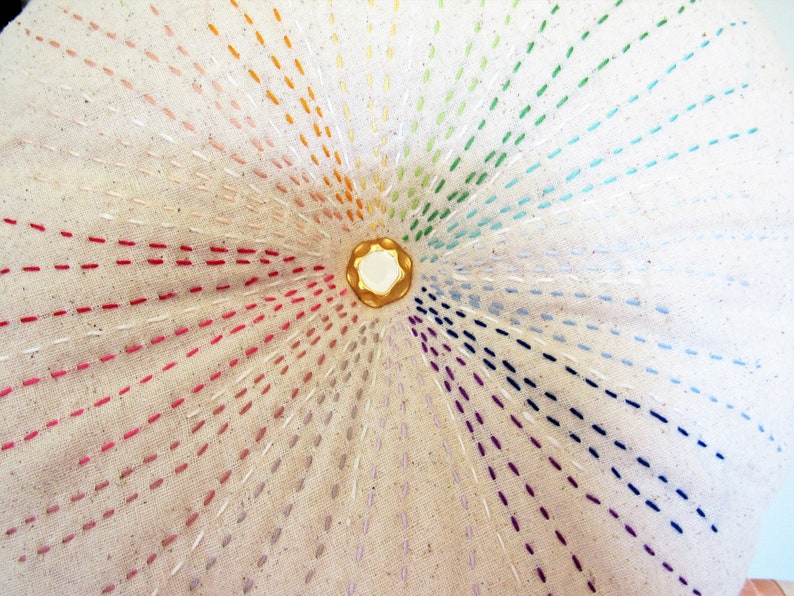 May include: A circular piece of fabric with a rainbow of colors stitched in a radial pattern around a gold button in the center.