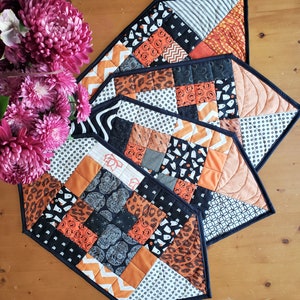 May include: Three black and orange patchwork table runners with a Halloween theme. The runners are made of different fabrics with various patterns, including polka dots, stripes, and skulls.