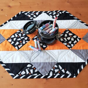 May include: A black and white quilted table runner with a black and white striped border. The runner is decorated with a pattern of white ghosts on a black background. A black jar filled with candy is on top of the runner.