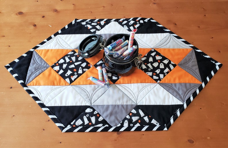 May include: A black and white quilted table runner with a black and white striped border. The runner is decorated with a pattern of white ghosts on a black background. A black jar filled with candy is on top of the runner.