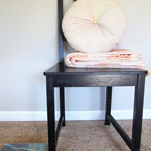 May include: A black wooden chair with a white round pillow with colorful stitching and a pink and white quilt folded on top.
