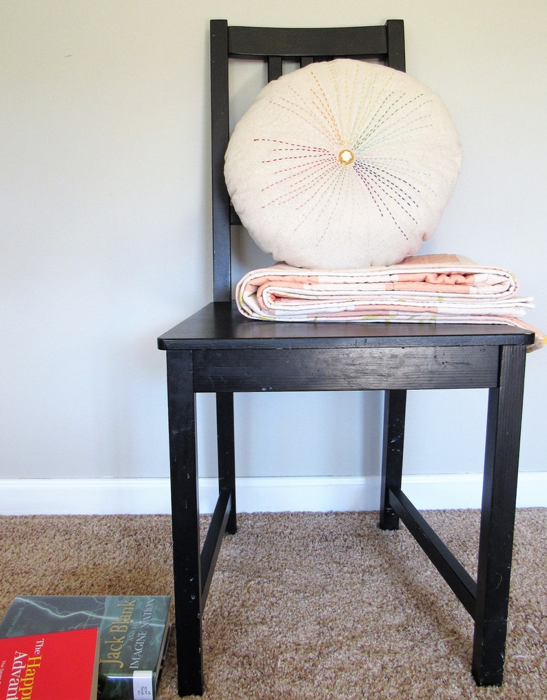 May include: A black wooden chair with a white round pillow with colorful stitching and a pink and white quilt folded on top.