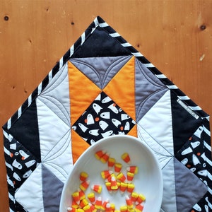 May include: A black, white, gray, and orange quilted table runner with a ghost pattern and a white bowl filled with candy corn.