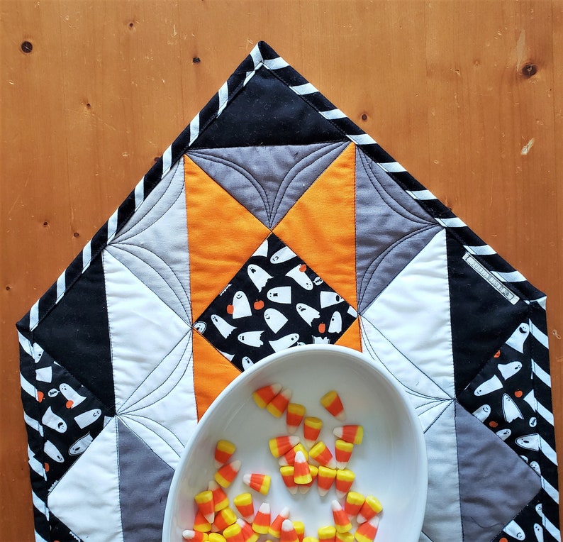 May include: A black, white, gray, and orange quilted table runner with a ghost pattern and a white bowl filled with candy corn.