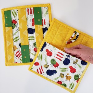 May include: Two yellow fabric pot holders with a white background and colourful vegetable designs. One pot holder has a patchwork design with green, red, and yellow squares. The other pot holder has a repeating pattern of vegetables, including tomatoes, peppers, onions, and aubergine.