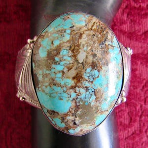 May include: A silver cuff bracelet with a large, oval turquoise stone with brown markings. The stone is set in a detailed silver setting. The bracelet is displayed on a dark surface against a red background.
