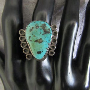 May include: A silver ring featuring a large, pear-shaped turquoise stone with dark brown matrix inclusions. The setting is accented by a series of small, circular silver wire loops arranged along both sides of the central gemstone.