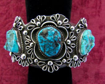 May include: A silver cuff bracelet featuring three turquoise stones. The bracelet has an elaborate floral design, with a central oval turquoise stone and two irregularly shaped turquoise stones. The bracelet is displayed against a black background.