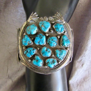 May include: A wide silver cuff bracelet featuring a large circular centerpiece adorned with twelve irregular, bright blue turquoise stones. The silver frame includes decorative metal frog-like accents at the top edge and small silver bead details between the stones.