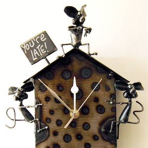 May include: A whimsical, handmade clock with a house-shaped frame. The clock face is brown with dark circular patterns. Metal mouse figures are perched on the clock, and one holds a sign that reads "You're LATE!" The clock hands are silver.