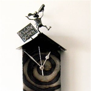 May include: A black metal clock with a swirling pattern on the face. The clock is shaped like a house with a small metal figure on top holding a bottle and a sign that says "It's wine o'clock!"