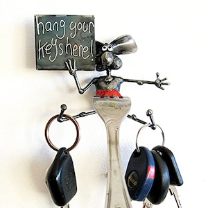 May include: A whimsical key holder made from metal, featuring a cartoon mouse holding a sign that says "hang your keys here!" The mouse is perched on a silver fork, and there are three key rings attached to the fork.