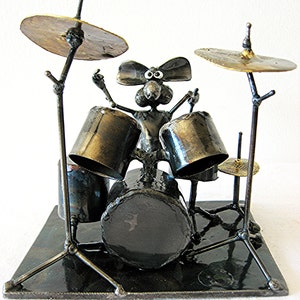 Könnte beinhalten: Eine Metallskulptur einer Maus, die ein Schlagzeug spielt. Die Maus ist aus Metall und hat große, runde Augen. Das Schlagzeug ist aus Metall und hat einen schwarzen Sockel. Die Skulptur ist ein skurriles und lustiges Kunstwerk.