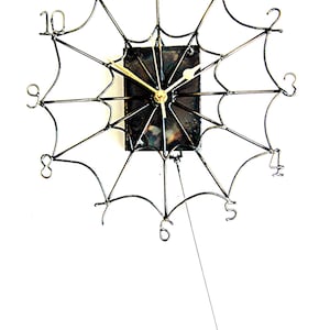 May include: A black metal clock shaped like a spider web with a black clock face and gold hands. A small metal spider hangs from the bottom of the clock.