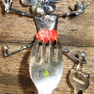 May include: A metal sculpture of a mouse holding a fork with a red piece of food on it. The mouse is holding the fork with its front paws and has its back legs extended out to the sides. The sculpture is attached to a key chain with a silver key.