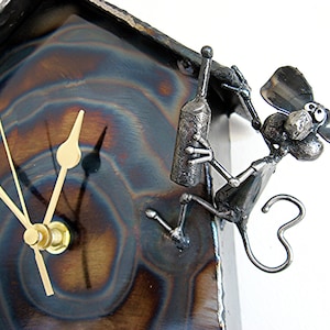 May include: A metal clock with a whimsical design featuring a small, metallic figure of a mouse holding a bottle. The clock face is surrounded by a textured, blue and brown metal surface.