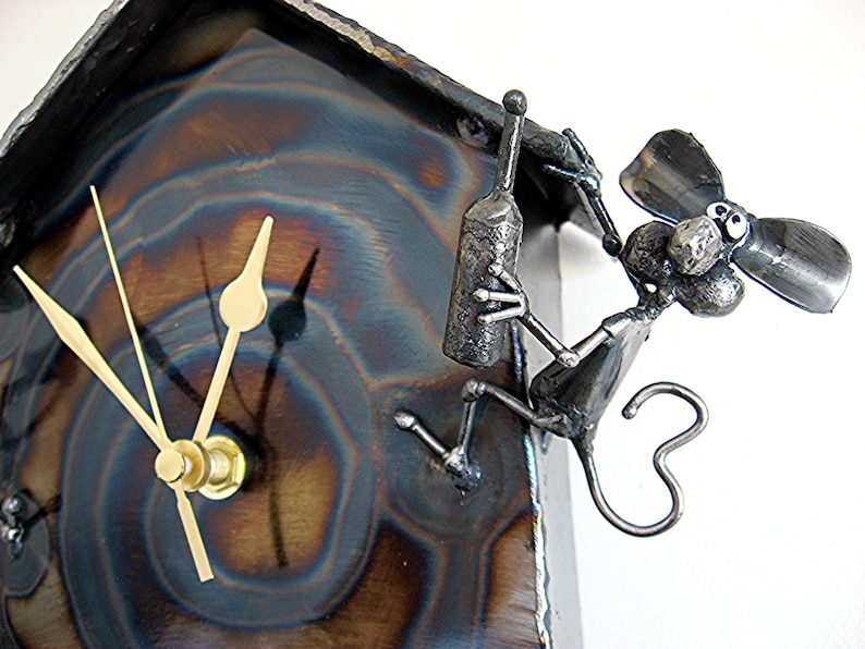 May include: A metal clock with a whimsical design featuring a small, metallic figure of a mouse holding a bottle. The clock face is surrounded by a textured, blue and brown metal surface.