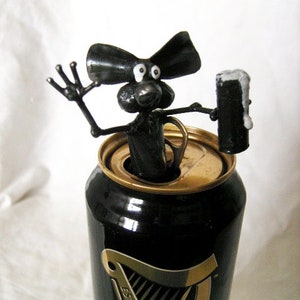 May include: A black metal sculpture of a mouse holding a can of Guinness beer. The mouse is sitting on top of the can, which is partially open. The mouse has a long tail and large ears.