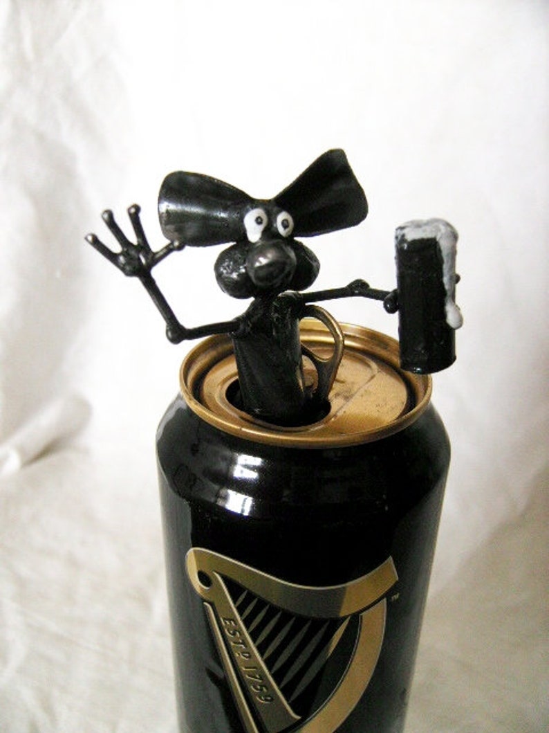 May include: A black metal sculpture of a mouse holding a can of Guinness beer. The mouse is sitting on top of the can, which is partially open. The mouse has a long tail and large ears.