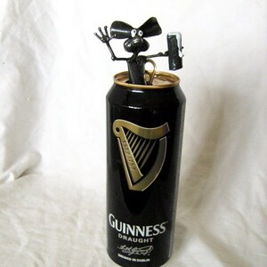 May include: A black metal figurine of a dog holding a can of Guinness Draught beer. The can is black with gold lettering and a harp logo. The text on the can reads "ESTD 1778" and "GUINNESS DRAUGHT".