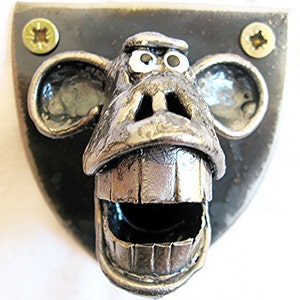 May include: A metal monkey face sculpture with a wide open mouth, large ears, and googly eyes. The sculpture is mounted on a black, shield-shaped metal plate and secured with two gold screws.